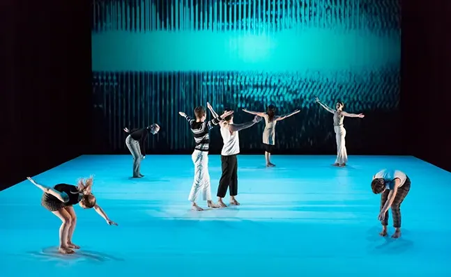 Seven dancers on stage against a bright blue floor and backdrop